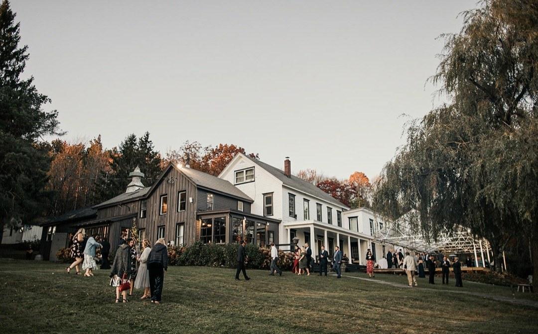 The Twelve Ponds farmhouse and barn at dusk, with guests gathered on the lawn under a string-lit tent.