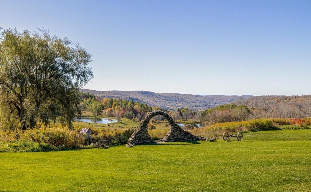 A stone arch sculpture on the meadow at Twelve Ponds, with autumn foliage and rolling hills behind.