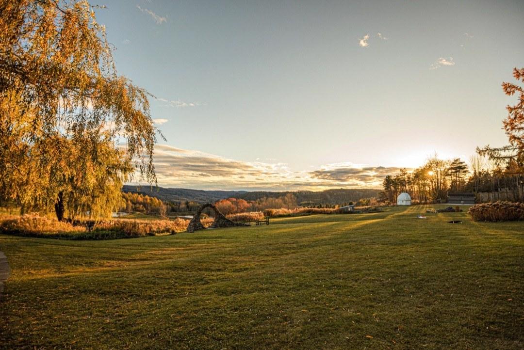 Meadow at golden hour with a willow tree, a stone arch sculpture, and the sun setting over the wooded hills in the distance.