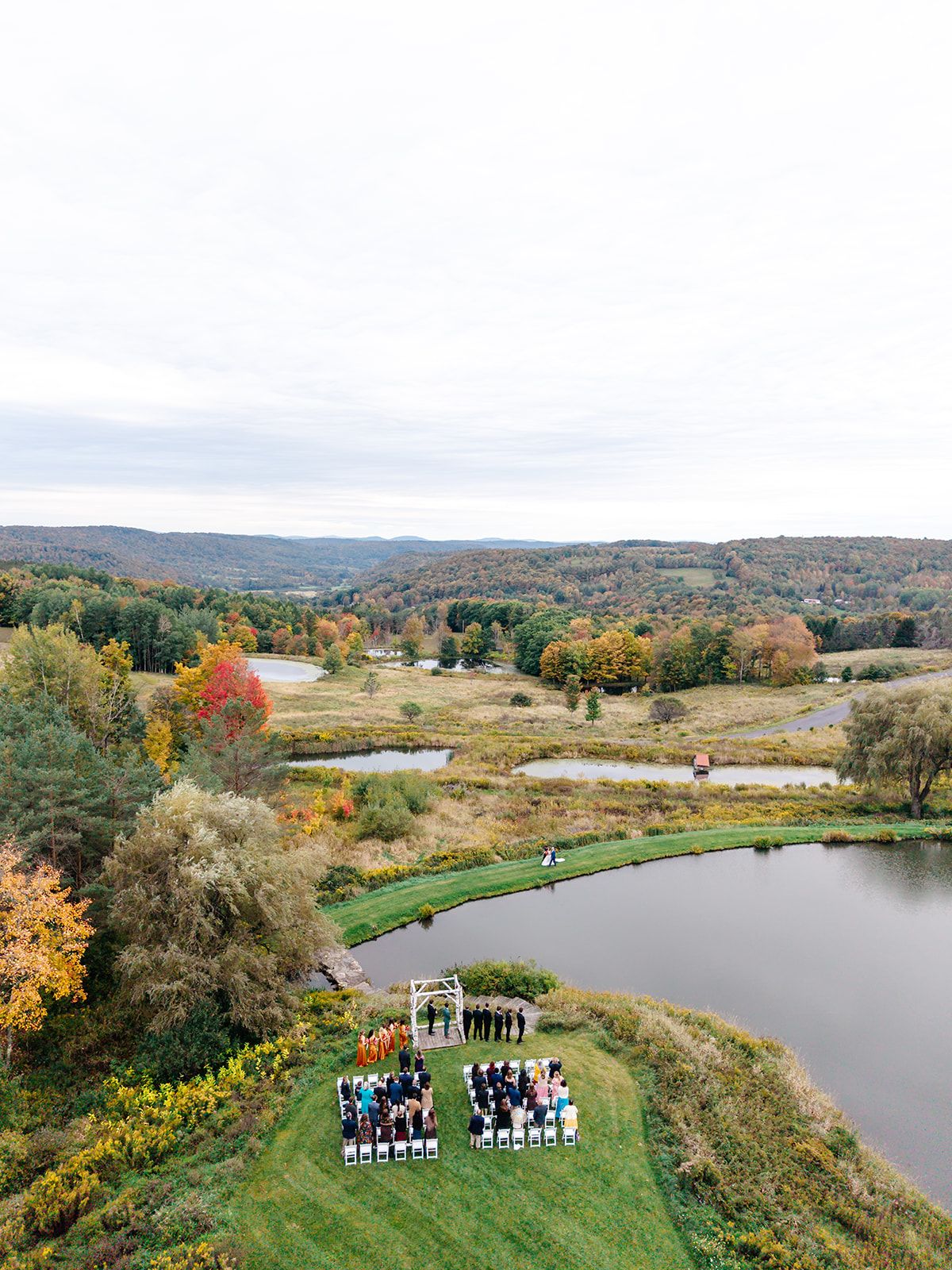 Aerial view of a wedding ceremony on the lawn beside a pond at Twelve Ponds, with autumn hills beyond.