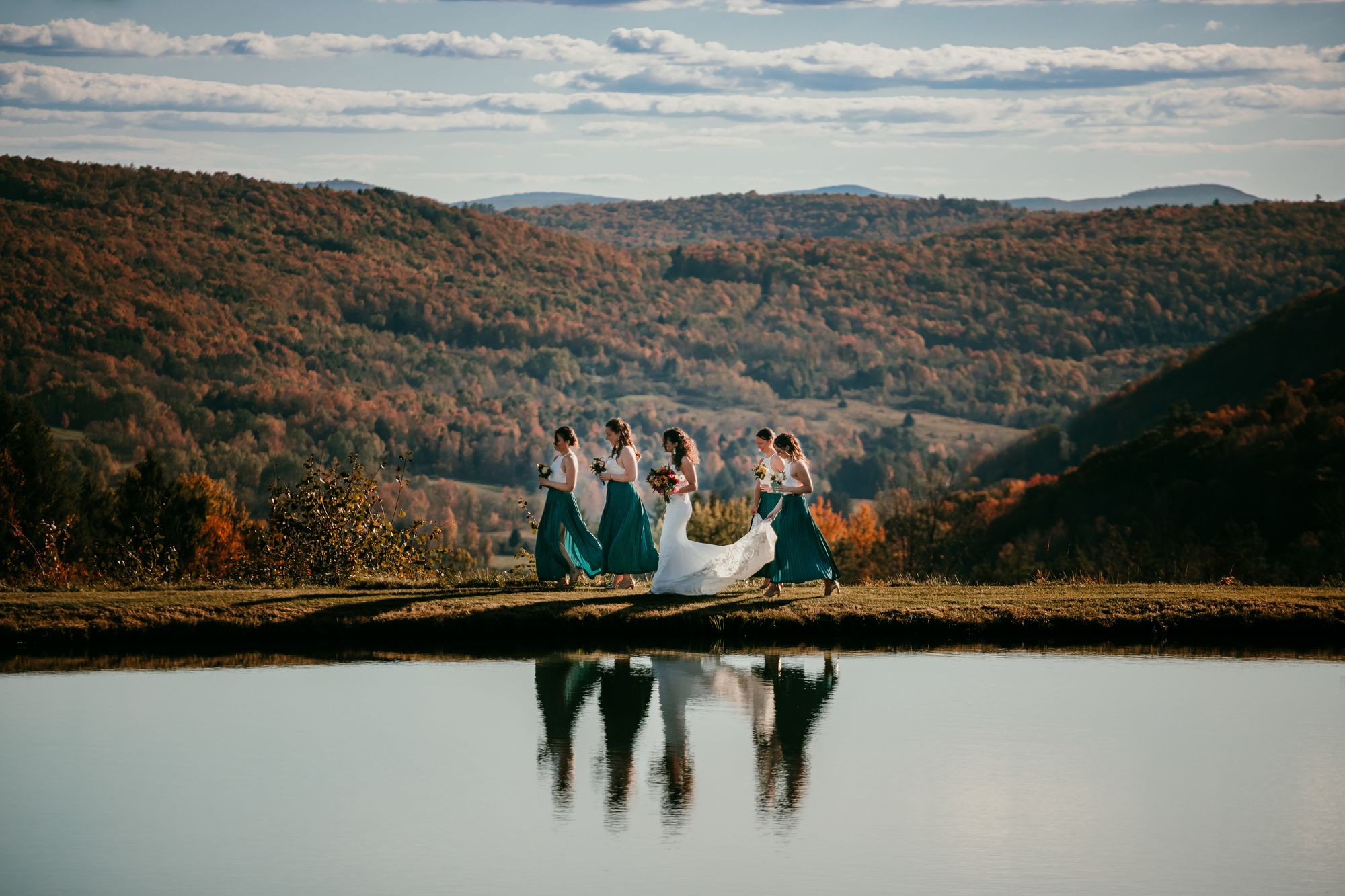 A bride and her bridesmaids walking along the edge of a pond at Twelve Ponds, reflected in the water, with autumn mountains behind.