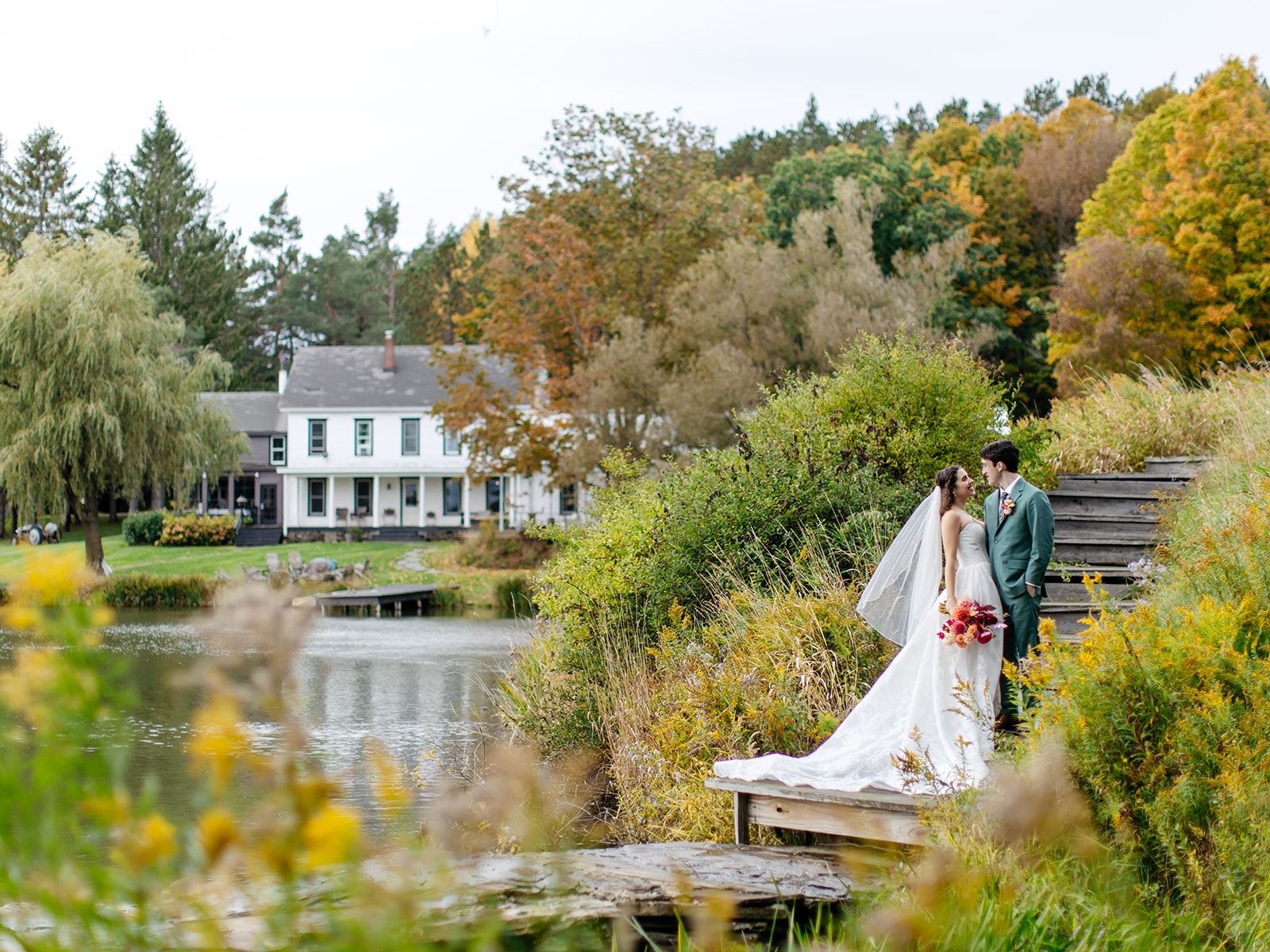 A bride and groom on a wooden dock at Twelve Ponds in autumn, with the farmhouse across the pond.