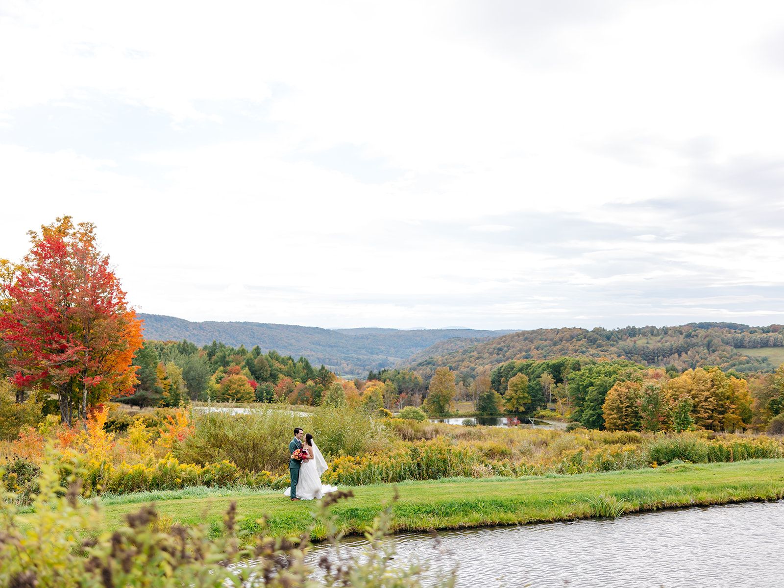 A bride and groom at the far edge of a pond at Twelve Ponds, with autumn hills beyond.