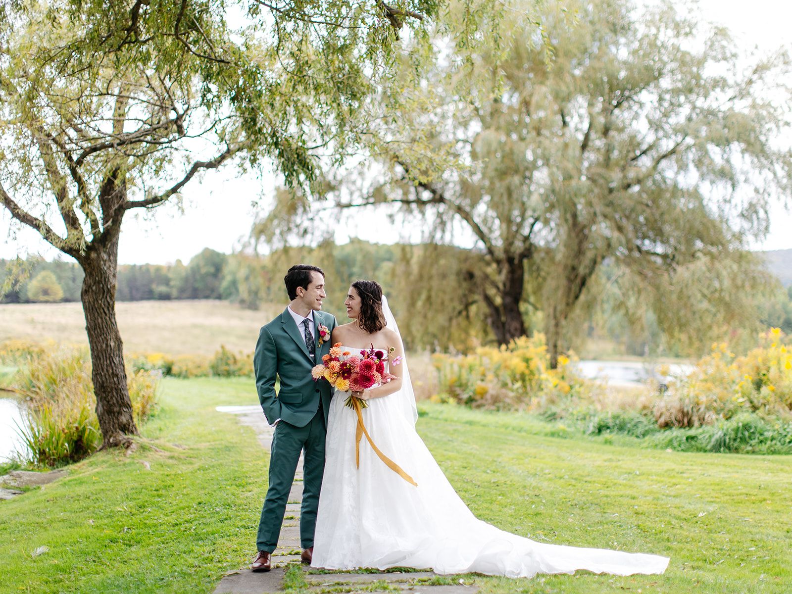 A bride and groom under a willow tree at Twelve Ponds, holding a bouquet of red and orange flowers.