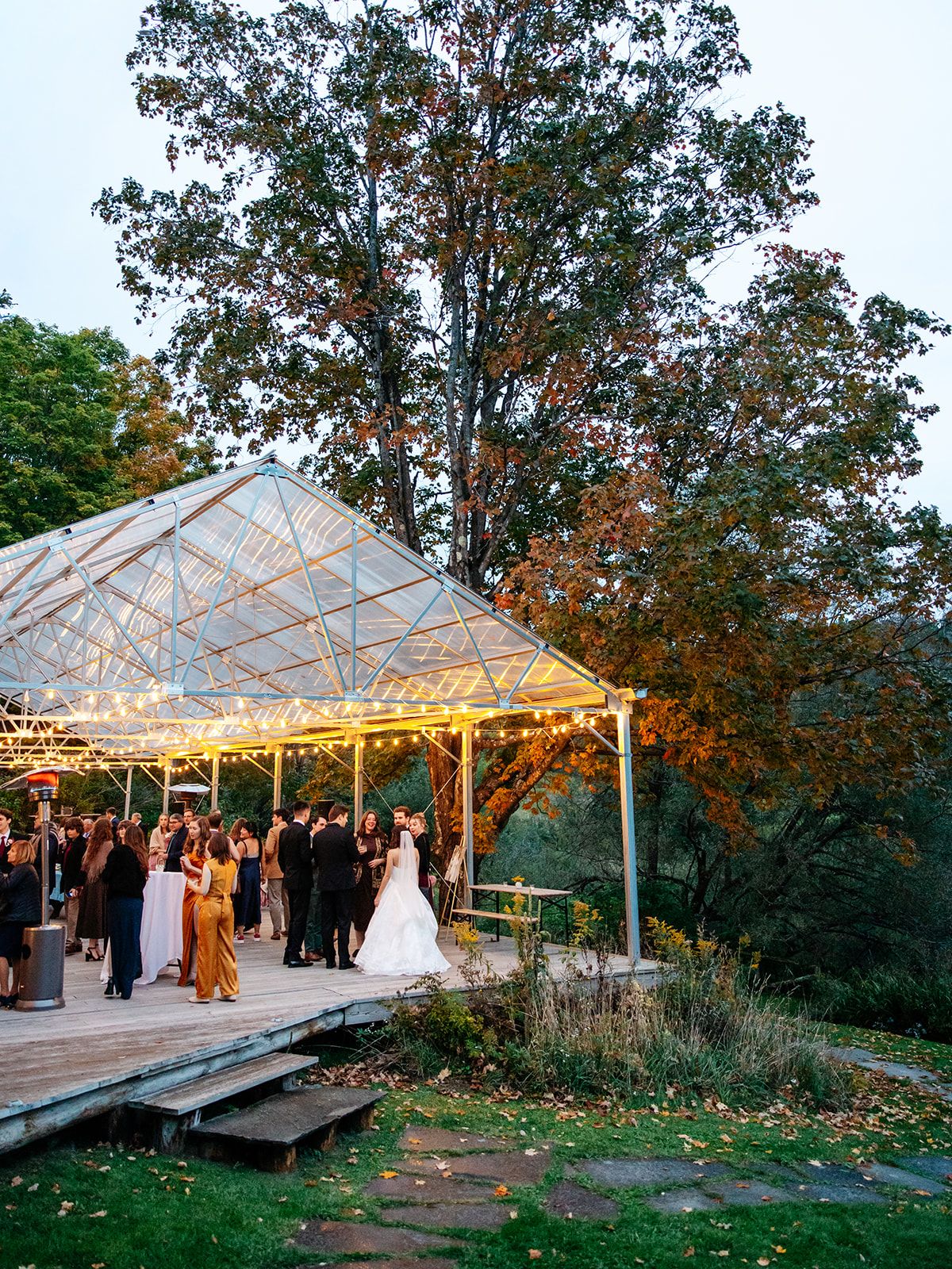 Guests at an evening reception under the glass pavilion at Twelve Ponds, lit with string lights among autumn trees.